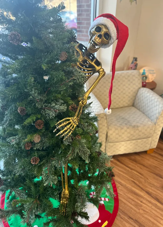 A skeleton wearing a long Santa hat peeking from behind a decorated Christmas tree