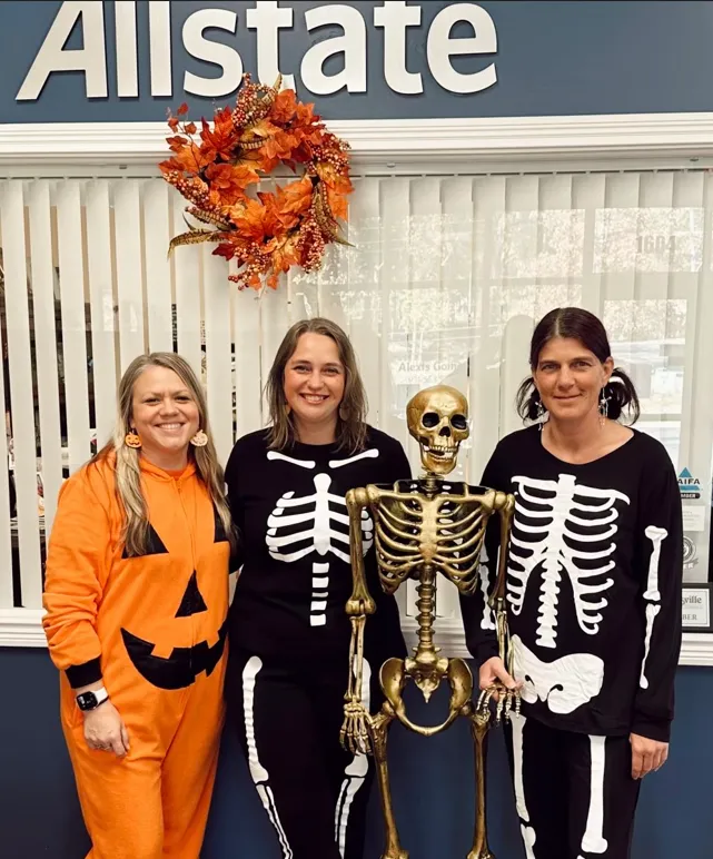 Three people in skeleton costumes standing with a skeleton beneath a large Allstate sign