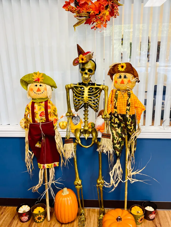 A gold skeleton wearing a pointed autumn hat sits between two scarecrow figures with pumpkins in an office window display