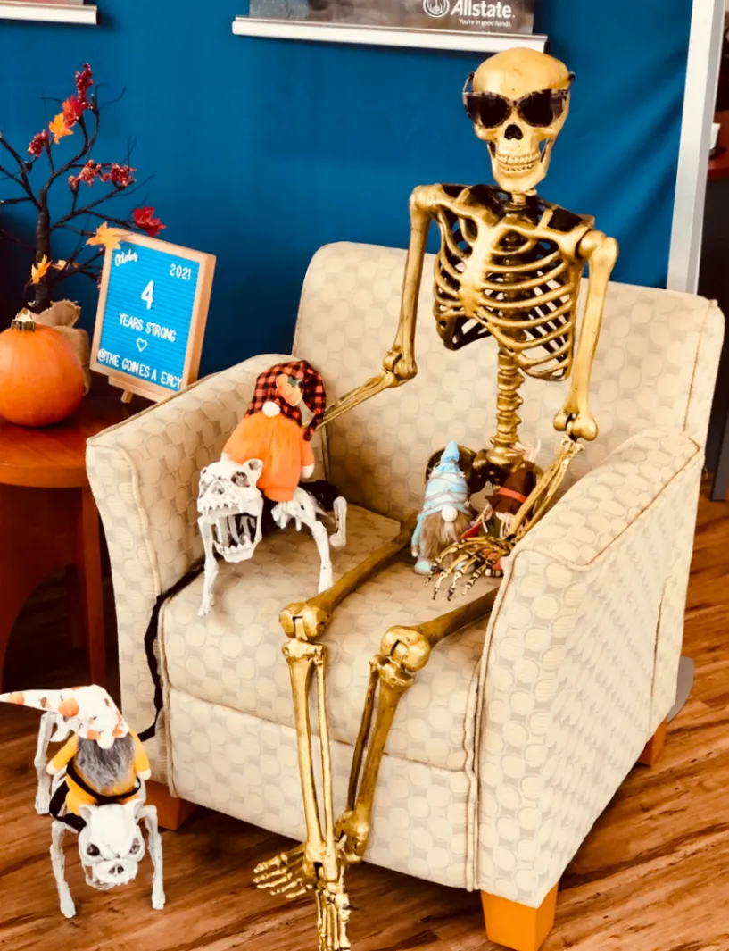 A skeleton wearing sunglasses sits in an armchair beside small skeleton figures and autumn decorations in an office