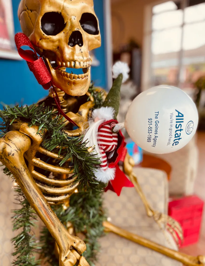 Close view of a skeleton holding a white Allstate ornament beside green garland and a holiday gnome