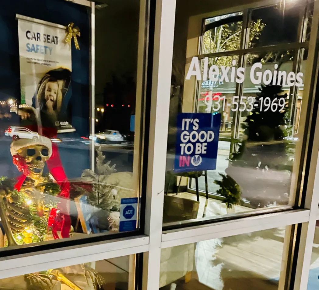 A nighttime view of an office window display with an illuminated skeleton, holiday decorations, and Alexis Goines lettering on the glass